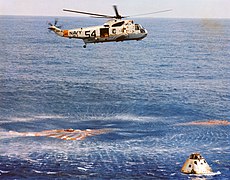 Photographie en couleur de la capsule amérisant avec l'hélicoptère de sauvetage au-dessus d'elle.