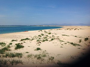 An empty crescent shaped beach, with small patches of grass and a small red boat sits on the shore. The sea appears to the left of the image and mountains appear in the background.