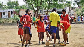 A group of young children standing around in dirt, soccer ball is at their feet and it looks as if they are discussing sport tactics.