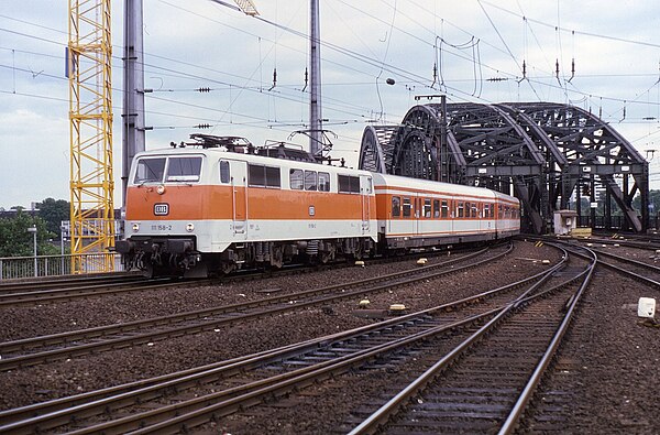 A Class 111 locomotive leads an orange-and-white S-Bahn service across the Hohenzollernbrücke into Köln Hauptbahnhof in 1985