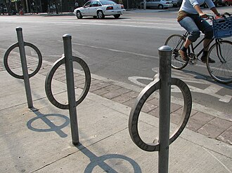 Неокрашенные стальные кольца, каждое приваренное к стальному столбу (проходящему через него), установленное в бетонных плитах. Велосипедист проезжает слева направо по велосипедной дорожке позади.