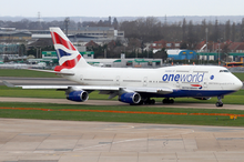 A British Airways Boeing 747-400 in Oneworld livery taxiing on the taxiway, with Heathrow Airport facilities in the background and green grass patch in the foreground