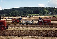 Brown field being ploughed by multiple red tractors. In the background it a hill with trees.