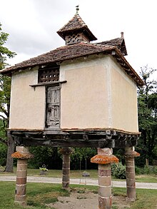 Farbfoto eines quadratischen Gebäudes mit einer Holzstruktur auf Steinsäulen.  Das Dach mit 4 Gefällen in flachen Ziegeln und oben von einem mit runden Löchern durchbohrten Holzgipfel überragt.