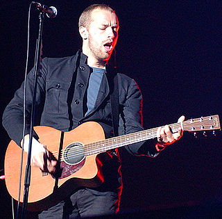 A man wearing a blue T-shirt and dark blue jacket holding a guitar and standing behind a microphone stand.