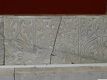 Een stenen blok volledig bedekt met houtsnijwerk van twee afwisselende bloemmotieven, gescheiden in vierkante delen