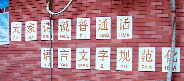 A school slogan asking elementary students to speak Standard Chinese is annotated with pinyin, but without tonal marks.