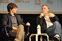 Photo de deux hommes assis devant un écran de projection d'une salle de cinéma. Celui de droite tient un micro dans sa main et parle en regardant en face de lui, tandis que l'homme de gauche l'écoute et le regarde.