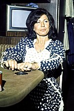 A black-haired woman in a dark blue outfit with white polka dots, looking contemplative while sitting at a table
