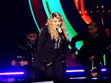 Madonna singing on a blue-lit stage in black costumes, surrounded by dancers. Behind them, there's a blue and orange backdrop