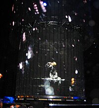 A circular stage on which a singer is seen on a piano. She is partially obscured by a circular screen covering her. The screen shows waterdrops falling.