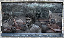 Metallrelief mit leichten, dreidimensionalen Elementen, Tom Pryce im Porträt mittig dargestellt, links und rechts zwei Shadow-Formel-1-Fahrzeuge, im Hintergrund eine Rennstrecke und eine Hügellandschaft mit Wäldern
