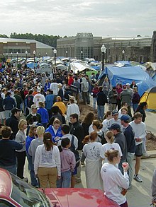 A large group of individuals gather in a parking lot alongside a tent campground with lightposts