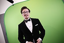 Smiling man in a black suit and bowtie, with a green background