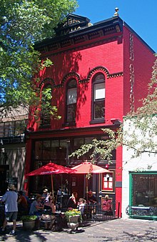 یک ساختمان باریک آجر قرمز سه طبقه با تزئینات مشکی تزئین شده ، از دو طرف به ساختمانهای پایین تر متصل است که از سمت راست آن دیده می شود ، شاخه های درخت از دو طرف به قاب وارد می شوند. کلمات 