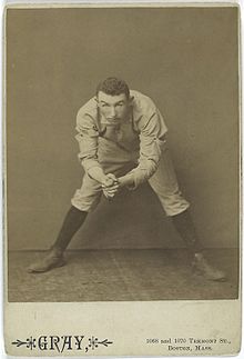 Ein Schwarzweißfoto eines Mannes in einer weißen Baseballuniform, der sich von der Taille gebeugt hat, aber in die Kamera schaut