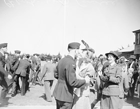 Muž v tmavé vojenské uniformě v náručí civilní ženy, mezi davem dalších civilních a vojenských lidí