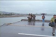 Scene from on the bridge post-collapse. Several reporters can be seen with umbrellas, and the weather is cloudy. The road is completely broken off and bent slightly upwards.