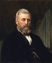 A portrait of a man on a dark background, wearing a white shirt mostly covered by a black suit jacket. He has mostly grey hair brushed back from a wide widow's peak, and a darker full beard and moustache with numerous grey spots. He is seated facing forward and to his left.
