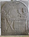 Stèle funéraire représentant la défunte assise buvant, devant une table où sont disposés les mets du banquet funéraire, avec un serviteur l'éventant. Zincirli, Pergamon Museum.