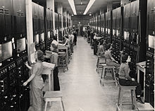 Two rows of control panels with dials and switches. Operators sit at them on four-legged stools.