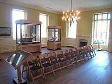 a yellow room with white molding and wainscoting. There is a row of chairs down the middle and two cases holding museum objects.