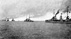 A long line of light gray warships bristling with guns steam in calm seas.
