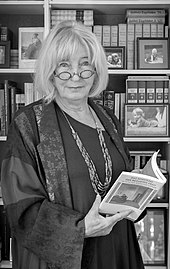 Zu sehen ist Inge Stephan stehend vor einem Bücherregal. Es ist eine schwarz-weiß Aufnahme. Sie blickt in die Kamera, hat schulterlanges Haar und trägt eine Lesebrille, welche ihr auf der Nase sitzt. Sie hält ein aufgeschlagenes Buch in der Hand.