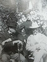Deux persommages assis avec chapeaux et une dame debout derrière les deux hommes.