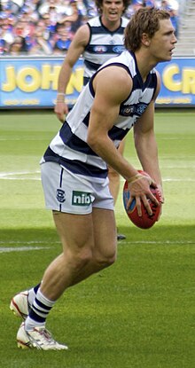 Le jeune athlète masculin se tient dans une chemise sans manches blanche et bleu marine et rayée et un short blanc.  Il se tient seul et regarde vers la droite tout en préparant le coup de pied au ballon.