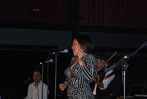 Upper body shot of a woman, singing and dancing before a microphone. She is shown in left profile and wearing a black and white dress. Behind her two male musicians are partly obscured or out of shot.