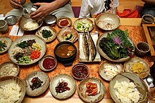 An array of about 10 small side dishes, a bean curd stew, and leaf vegetables on a table.