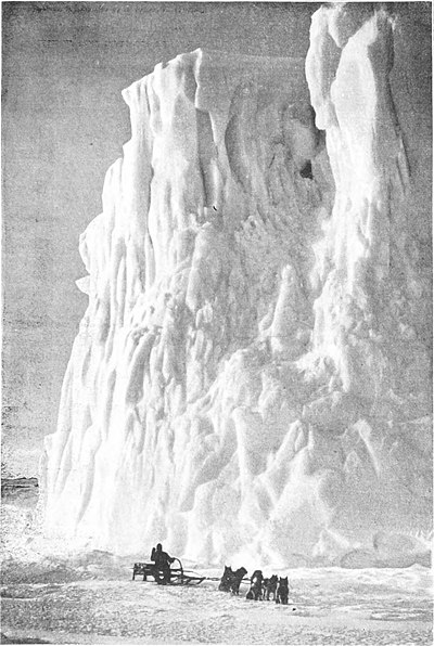 Photo of a rugged ice cliff dwarfing a dogsled team at its foot