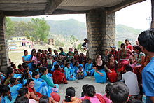 Sous un abri en pierre et en bois, assemblée de jeunes filles népalaises assises par terre en tenue bleue, assistant à un cours de sensibilisation.
