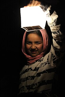 Menina refugiada síria de 14 anos à noite, vestida com um lenço na cabeça, é iluminada por uma lanterna solar chamada SolarPuff no campo de refugiados de Idomeni, na Grécia 2015