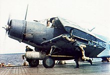 Barevná fotografie jednomotorového bombardéru pojíždějícího po palubě letadlové lodi. Letoun má sklopená křídla.