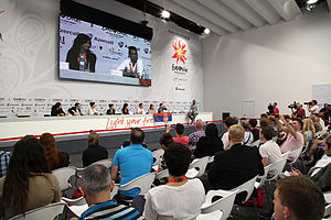 A press conference during the 2012 contest; the Serbian delegation are seated at a long table with rows of journalists seated facing them, with a large screen on the wall behind the delegation projecting a live relay of the conference.