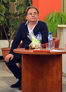 Na fotografii widoczny jest Robert Makłowicz siedzący przy małym drewnianym okrągłym stole. W tle rośliny doniczkowe