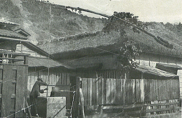 A man at a well, alongside buildings with the thatched roofs, weather-beaten unpainted sides and paper partitions and windows, characteristic of the i