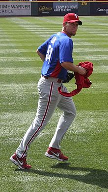 A man in grey pants, a blue baseball jersey, and a red baseball cap with 