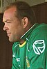 A man with receding, short brown hair is pictured from the side. He is wearing a green cricket uniform with the outline of a white flag in a shield predominantly on view on the shoulder.