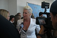 Mia Michaels, gekleed in een wit pak met bleekblond haar, staat voor een journalist terwijl ze wordt gefilmd door een onbekende cameraman.