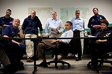 Grupa mężczyzn siedzi przy stole patrząc poza ekran, Obama w środku.