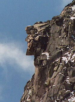 A jagged formation of granite rocks on a lightly snowed cliff forming the side profile resembling a human face juts out against the backdrop of a blue sky with a thin cloud to the left.
