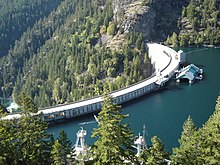 Un barrage en béton incurvé, vu d'un peu au-dessus.  Sur la gauche se trouve une vallée boisée escarpée et sur la droite, une eau bleu-vert.  Le réservoir est presque plein.