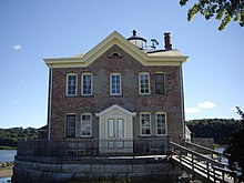 A brick building with yellow wooden trim and a pointed roof with a chimney and weather instruments on the roof. On the other side part of a lighthouse is visible. A wooden ramp leads up to a deck on its stone foundation surrounded by an iron fence.