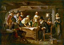 A group of men in antiquated clothing gathered around a desk in a ship's cabin with one woman looking on. A large piece of paper is on the desk and one man is signing.