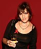 Tracey Emin, sosteniendo una copa de vino llena de un líquido translúcido de color melocotón.  Lleva un top negro, con una cinta roja adjunta.