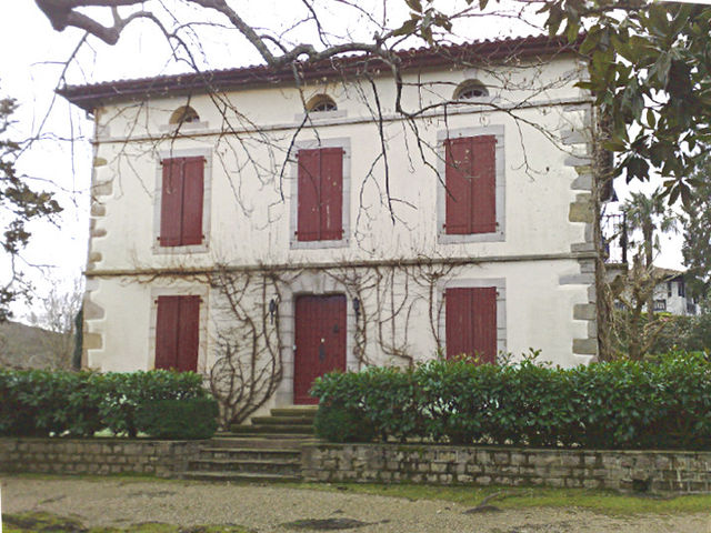 Photographie d’une maison à un étage, aux volets rouge sang clos.