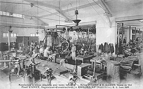 Photo noir et blanc de l'intérieur d'une usine avec des ouvriers, des machines outils et des courroies de transmission.